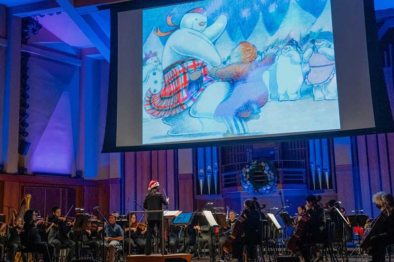 On a large screen a snowman dances around in the snow with a young boy. Below the screen is a symphony playing.