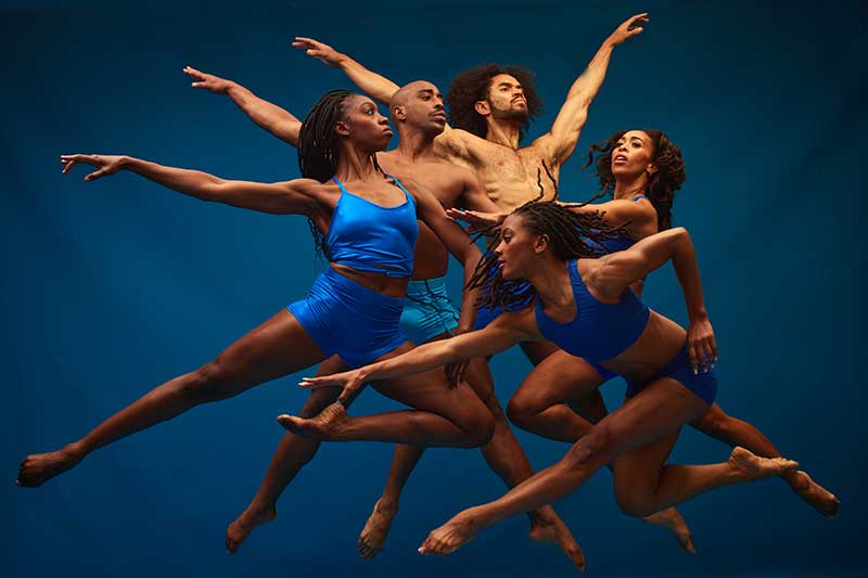 A group of Black dancers wear blue and leap together with arms extended.