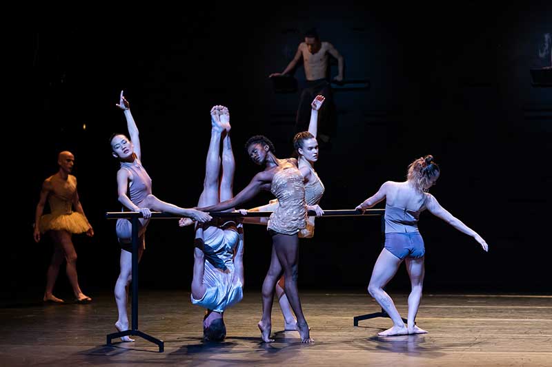 A dance company stands on stage. They hold a ballet bar in varying poses. One dancer is upside down on their head.