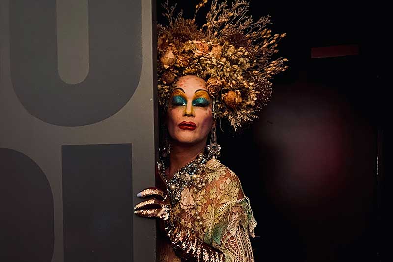 Performer Taylor Mac wears a grand headdress and gold dress while standing partially behind a wall