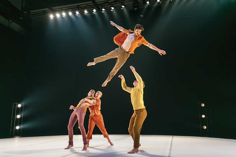 A group of dancers. One is high up in the air leaping from two dancers to a third.