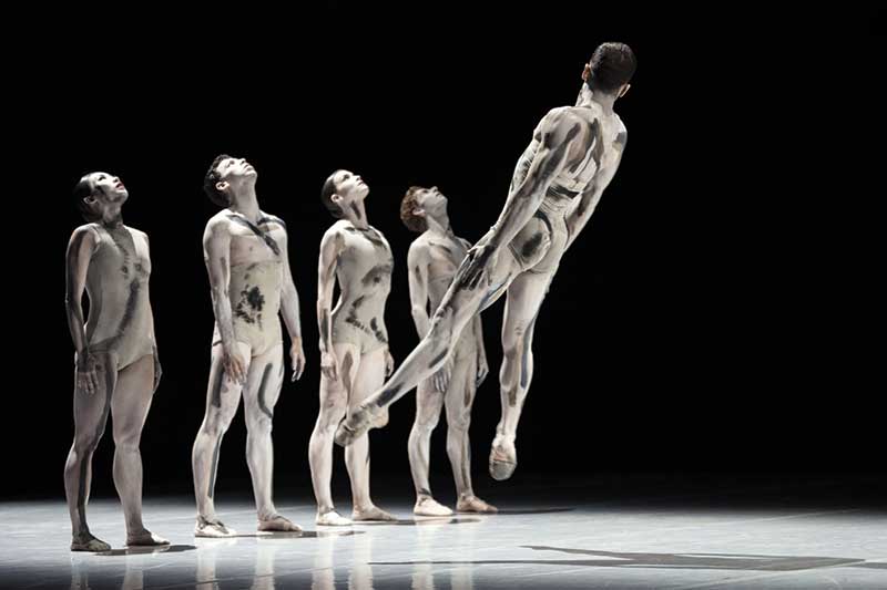 A group of dancers wearing white and white paint stand with their heads looking straight up while another dancer leaps backwards.