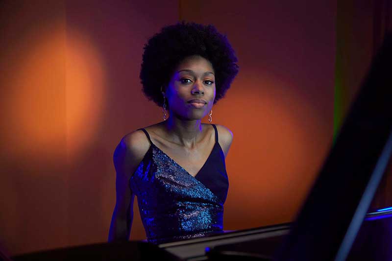 A young black woman wearing a blue tank top sits behind a piano in front of an orange background.