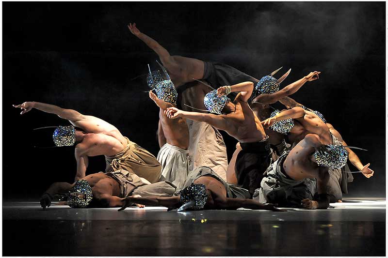 A group of dancers have their arms outreached. They wear reflective masks.