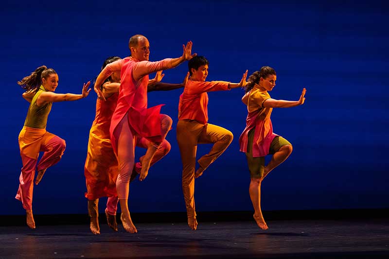 A group of dancers wearing orange move in synchronization in front of a blue background.