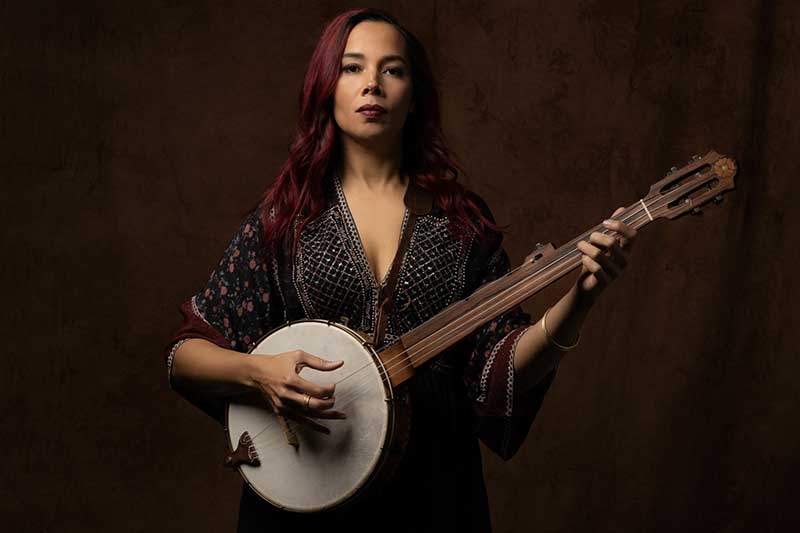 A white woman with red hair stands in front of a dark background holding a banjo.