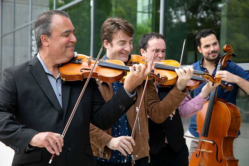 Four white men ranging in age, stand in a row playing string instruments.