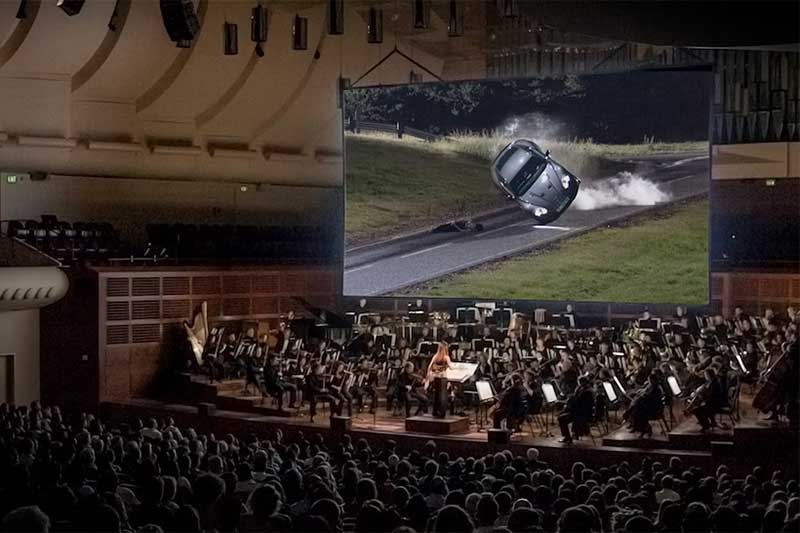 A crowd of people watch a James Bond movie on a big screen with an orchestra playing below.