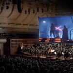 A crowd of people watch Star Wars on a big screen with an orchestra playing below.