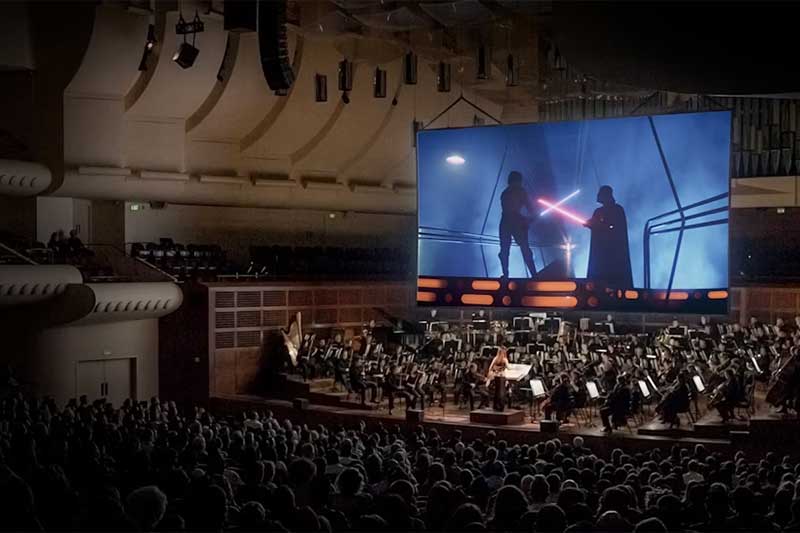 A crowd of people watch Star Wars on a big screen with an orchestra playing below.