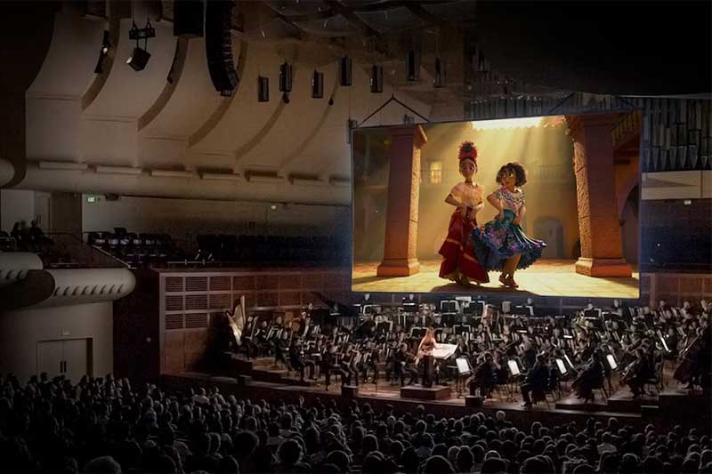 A crowd of people watch Encanto on a big screen with an orchestra playing below.