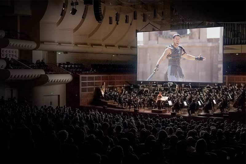 A crowd of people watch Gladiator on a big screen with an orchestra playing below.