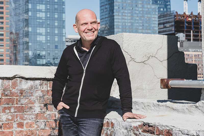 A white bald man stands on a rooftop in a city smiling.
