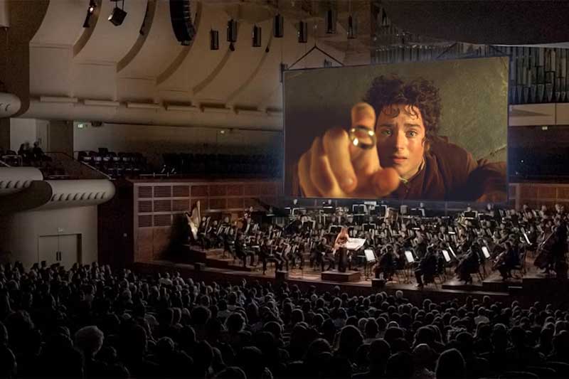 A crowd of people watch Lord of the Rings on a big screen with an orchestra playing below.