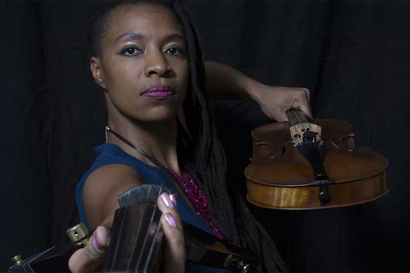 A black woman with brightly colored makeup stands in front of a black background and holds a violin almost like a bow and arrow.
