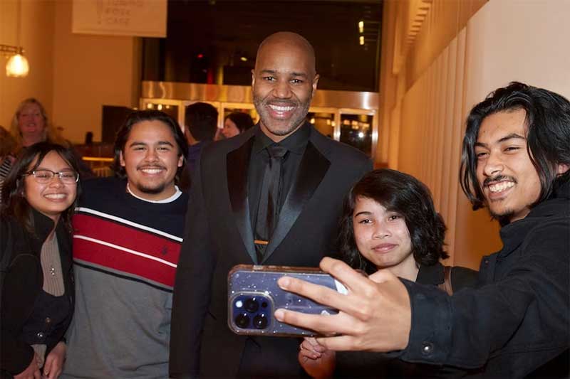 A group of teens stands with an older man as they take a selfie.
