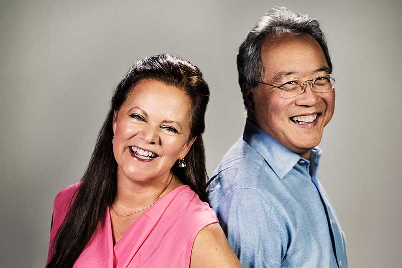 A middle aged woman and man stand back to back smiling.