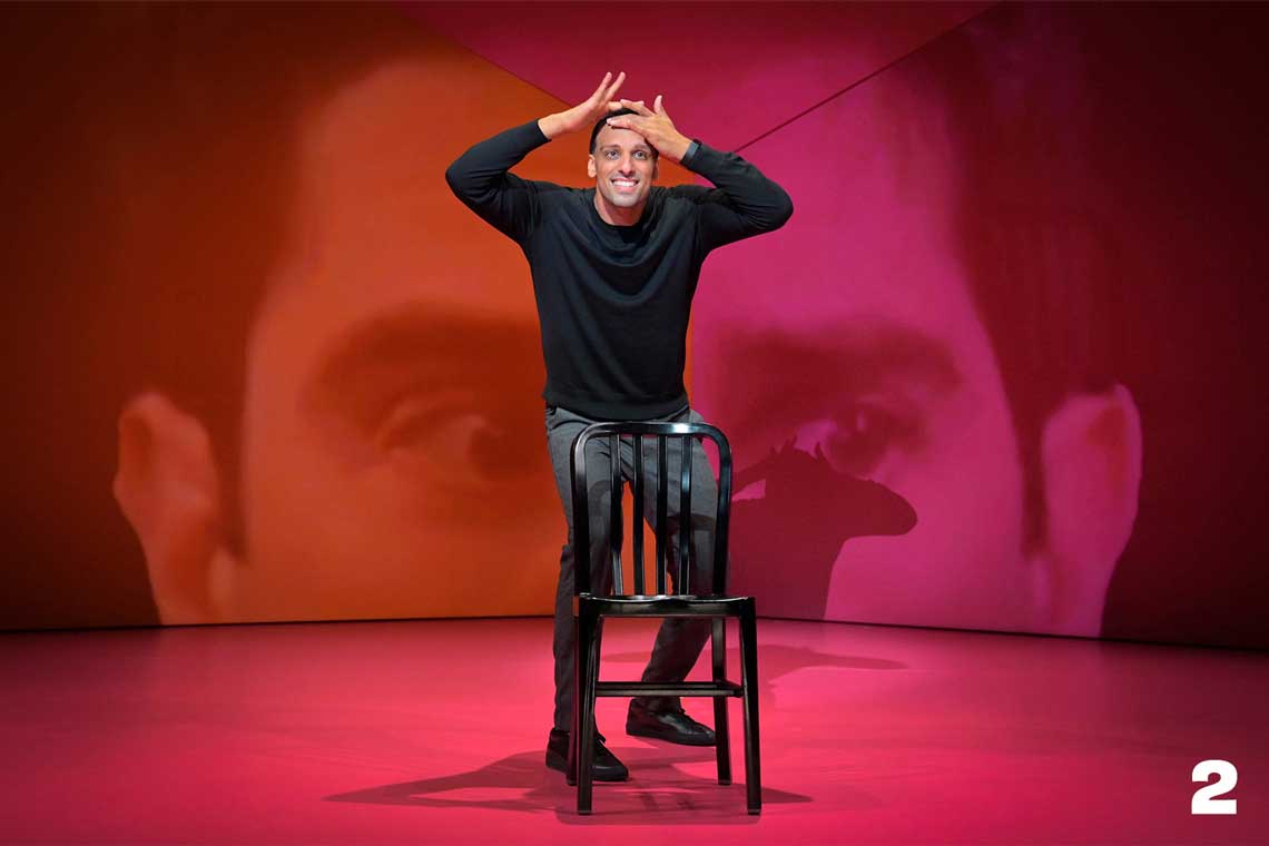 A man in a black shirt stands behind a stool with a pink and orange background.