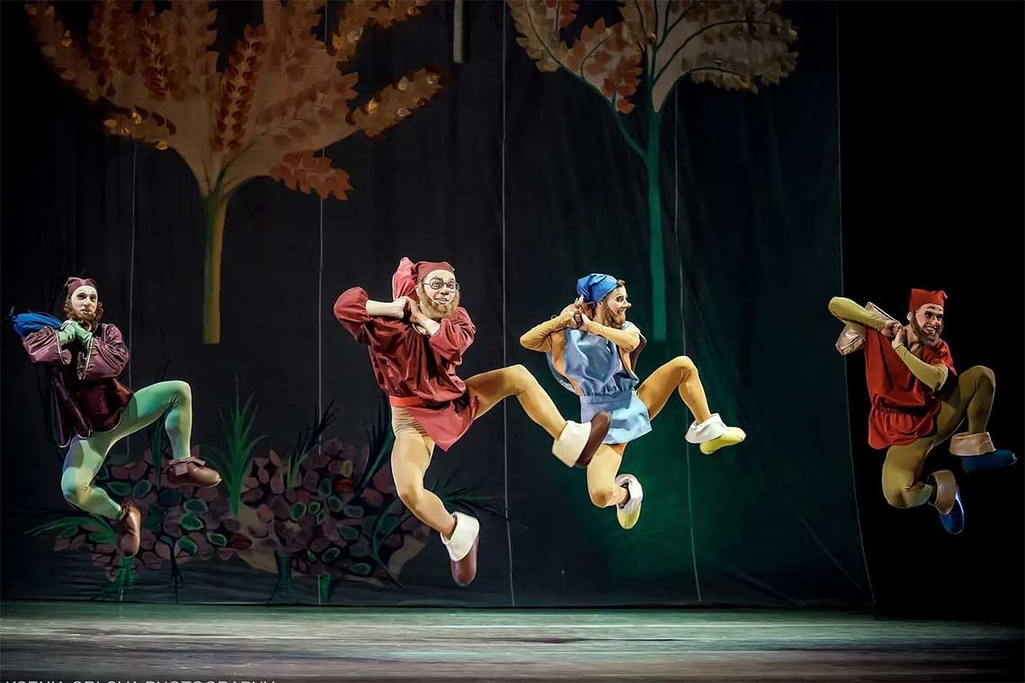 On a stage, four men dressed as the dwarves from snow white are leaping in the air in a dance.