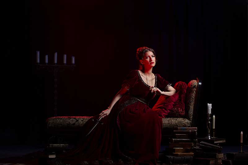 A white woman wearing a deep red dress sits on a couch.