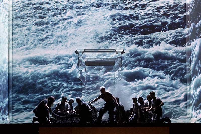 A large stage background shows waves on a sea. In the foreground are a group of men.