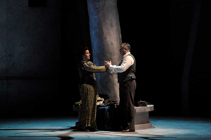 A man and woman stand on either side of a large tree and hold hands, encircling the tree.