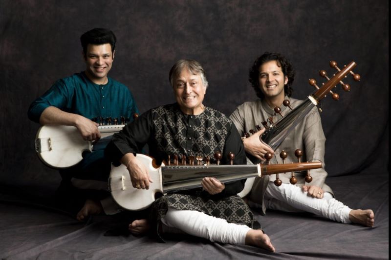A group of three men sit on the ground holding banjos