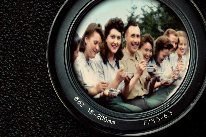 Within the reflection of a camera lens a group of young people smile, dressed from the 1940s.