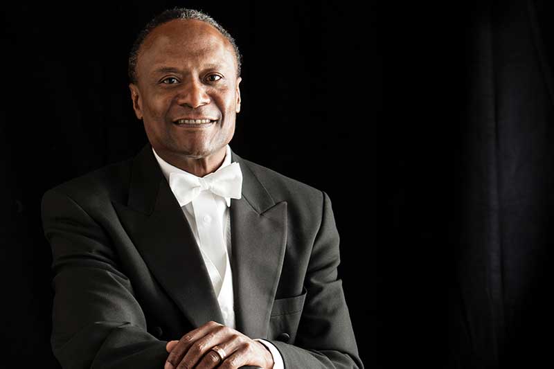Conductor Thomas Wilkins sits in a tuxedo smiling.