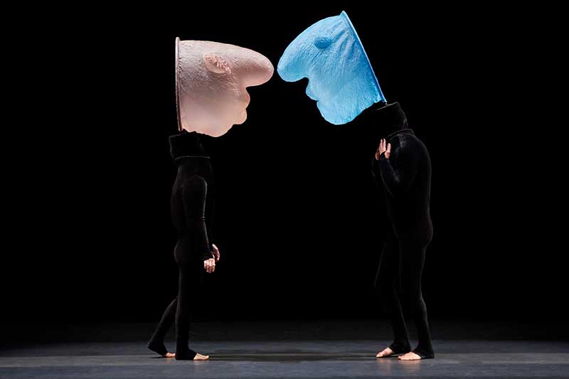 Dancers of Mummenschanz on stage wear black outfites with large facelike masks above their heads