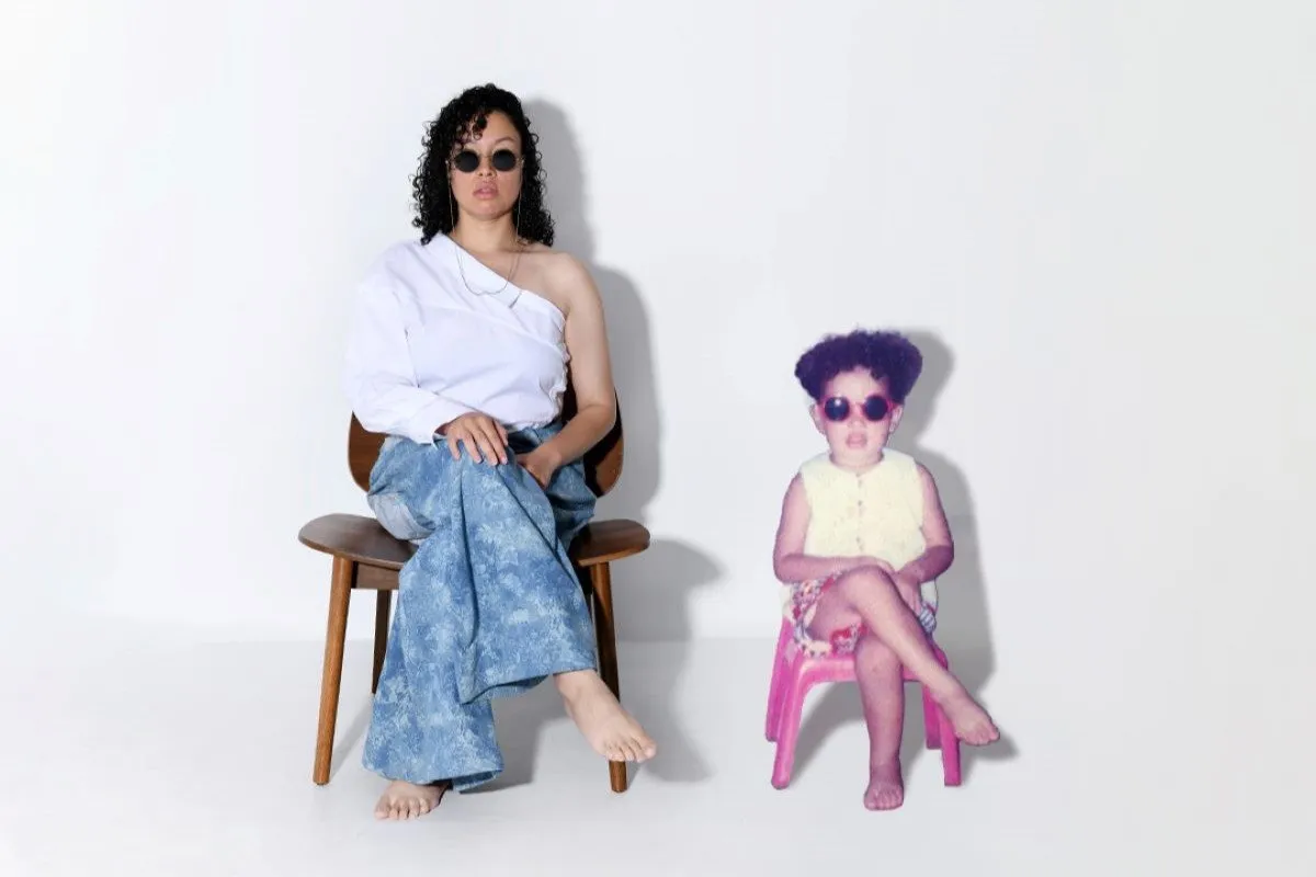 A woman and child sit in front of a white stark background. They both look at the camera wearing sunglasses.