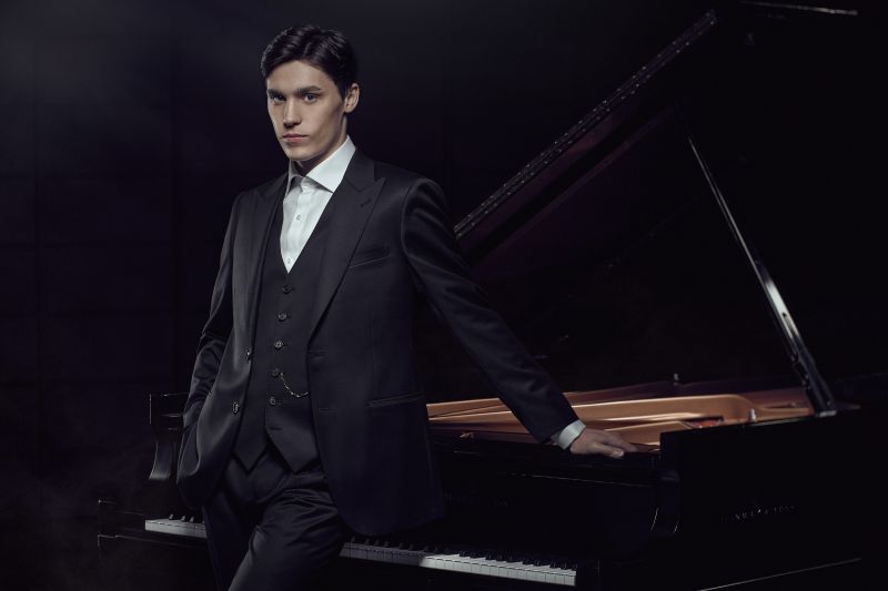 Jaeden Izik Dzurko wears a suit and stands next to a grand piano.
